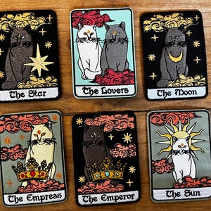May include: Set of ten embroidered iron-on patches featuring tarot card imagery with cats. Each patch features a different tarot card, including The Star, The Lovers, The Moon, The Empress, The Emperor, and The Sun. The patches are all black and white with colorful accents.