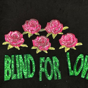 May include: A black fabric patch with the words "BLIND FOR LOVE" in green sequins. Five pink roses with green leaves are sewn above the text.