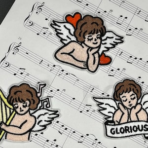 May include: Three embroidered iron-on patches featuring angels with wings. The first angel is holding a heart, the second is playing a harp, and the third is holding a banner that says "GLORIOUS".