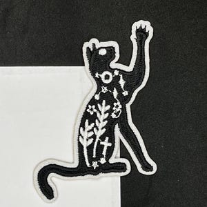 May include: Black embroidered iron-on patch of a cat with white floral and star designs.