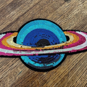 May include: A colorful sequined patch in the shape of a planet. The planet features a blue and black center with rainbow-colored rings of sequins. The patch is set against a black background.