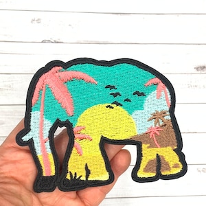 May include: An embroidered iron-on patch featuring a silhouette of an elephant with a tropical sunset scene inside. The elephant is outlined in black and filled with a turquoise blue, yellow, and pink color scheme. The scene includes palm trees, a setting sun, and clouds.