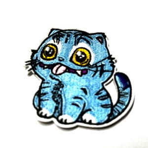 May include: Embroidered patch featuring a cartoon cat with blue fur, large yellow eyes, and a pink tongue sticking out. The cat has a white outline and is sitting with its tail curled.