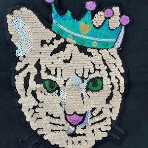 May include: A white tiger face with green eyes and a pink tongue, made of iridescent sequins, wearing a turquoise crown with a gold heart on top. The crown is also made of sequins.