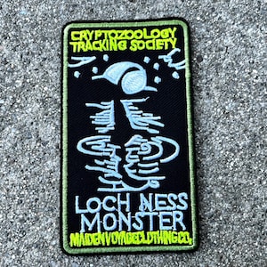 May include: A black embroidered patch with green stitching. The patch features a white outline of a creature with a long neck and a small head. The text "CRYPTOZOOLOGY TRACKING SOCIETY" is at the top of the patch, and the text "LOCH NESS MONSTER" is at the bottom. The text "MAIDEN VOYAGE CLOTHING CO." is at the bottom of the patch.