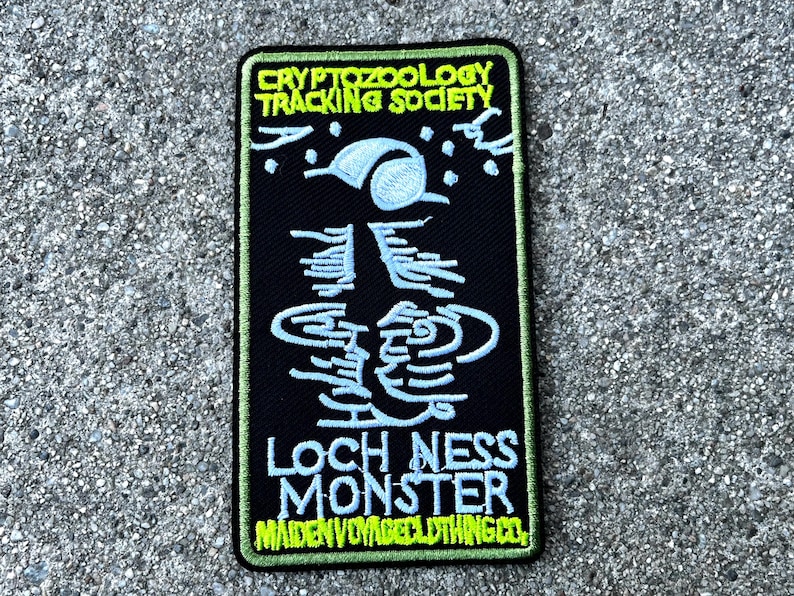 May include: A black embroidered patch with green stitching. The patch features a white outline of a creature with a long neck and a small head. The text "CRYPTOZOOLOGY TRACKING SOCIETY" is at the top of the patch, and the text "LOCH NESS MONSTER" is at the bottom. The text "MAIDEN VOYAGE CLOTHING CO." is at the bottom of the patch.