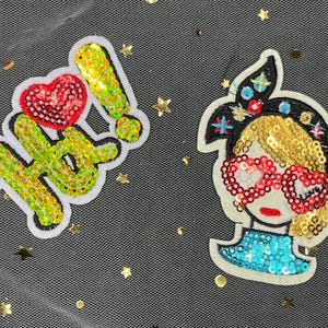 May include: Two iron-on patches with a sparkly, sequined design. One patch says "Hi!" with a red heart. The other patch features a cartoon girl with a blue headband, gold hair, and red heart-shaped sunglasses.