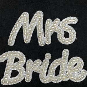 May include: White beaded "Mrs" and "Bride" lettering on a black background. The letters are outlined with small, clear rhinestones and filled with round, white beads. Ideal for bridal accessories.