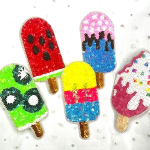 May include: Five colorful sequined popsicle patches. The popsicles feature various designs, including a watermelon, kiwi, and other fruit-inspired patterns. Each patch has a wooden stick and is embellished with sequins in vibrant colors.
