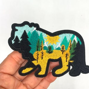 May include: An embroidered iron-on patch featuring a yellow lion silhouette with a forest scene inside. The forest scene includes green trees, blue mountains, and a yellow sun.