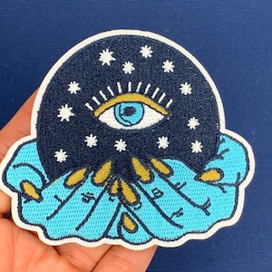 May include: Embroidered patch with a blue eye surrounded by white stars on a dark blue background. The eye is surrounded by a gold-colored eyelid. The patch is held by two blue hands with gold accents.