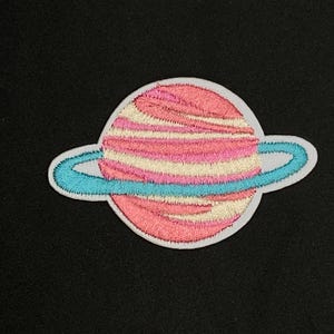 May include: A pink and white striped planet with a blue ring around it. The planet is embroidered on a white fabric.