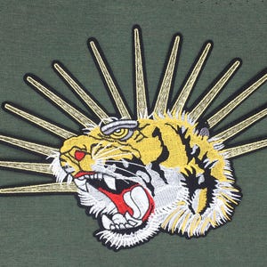 May include: Embroidered tiger head with a yellow and white striped pattern, with a red mouth and white teeth, surrounded by a golden sunburst on a green background.