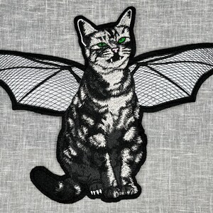 May include: A black and white cat with green eyes and a black bat-like wing design on its back. The cat is sitting upright with its wings spread out.