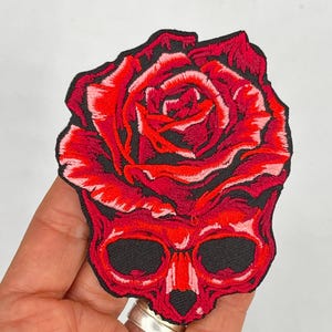 May include: Red embroidered patch of a skull with a red rose growing out of the top of its head.