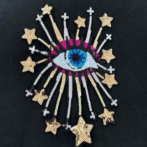 May include: A blue eye with pink and white accents surrounded by gold and silver sequins in the shape of stars. The eye is surrounded by a burst of gold and silver sequins.