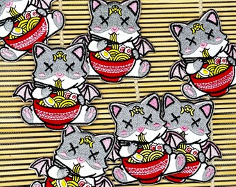 May include: Embroidered patches featuring a gray cat with bat wings, eating ramen from a red bowl. The cat has X-shaped eyes and a crescent moon and star on its forehead. The patches are on a bamboo mat.