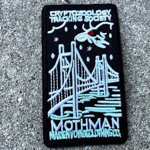 May include: Black embroidered patch with white and blue stitching. The patch features a bridge, a mothman, and the text "Cryptozoology Tracking Society" and "Mothman Maiden Voyage Clothing Co."