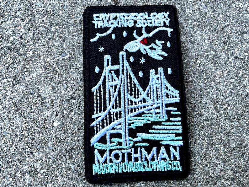 May include: Black embroidered patch with white and blue stitching. The patch features a bridge, a mothman, and the text "Cryptozoology Tracking Society" and "Mothman Maiden Voyage Clothing Co."