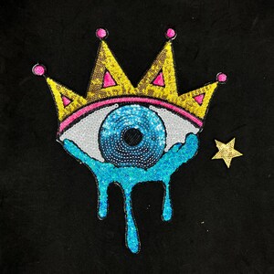 May include: A blue eye with a crown on top and dripping blue tears. The eye is surrounded by sequins and the crown is yellow with pink accents. A gold star is in the background.