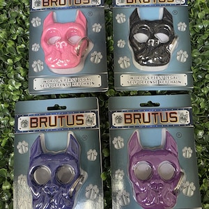 May include: Four "Brutus" self-defense keychains in pink, black, purple, and lavender. Each keychain is shaped like a dog's face and includes a keyring. The packaging reads "World's First Legal Self Defense Keychain."