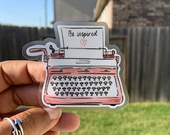 May include: A pink typewriter sticker with the text "Be inspired" and a small heart above the text. The sticker is on a clear background.