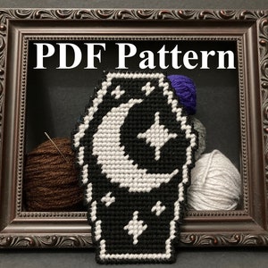 May include: A black and white cross-stitch pattern of a coffin with a crescent moon and stars. The pattern is framed in a dark brown frame with the text "PDF Pattern" above it.