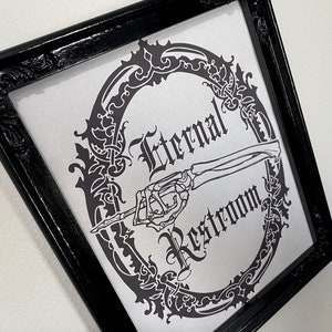 May include: A black framed sign with a white background and a black and white illustration of a skeleton hand pointing to the right. The sign reads "Eternal Restroom" in a gothic font.