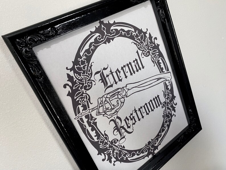 May include: A black framed sign with a white background and a black and white illustration of a skeleton hand pointing to the right. The sign reads "Eternal Restroom" in a gothic font.