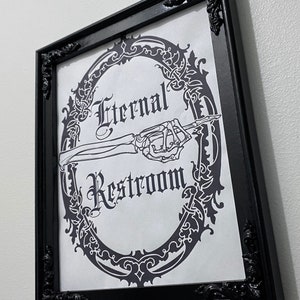 May include: Black and white sign with an ornate frame and a skeleton hand pointing to the right. The sign reads "Eternal Restroom".