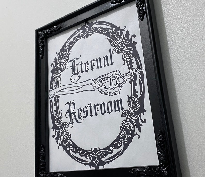 May include: Black and white sign with an ornate frame and a skeleton hand pointing to the right. The sign reads "Eternal Restroom".