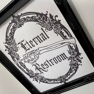 May include: A black framed print with a white background. The print features a skeletal hand pointing to the right with the text "Eternal Restroom" in a gothic font.