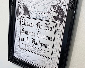 May include: A black framed sign with a white background and a spiderweb design. The sign reads "Please Do Not Summon Demons in the Bathroom. Spirits in mirror are not as charming as they appear."