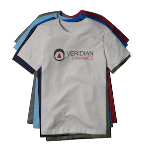May include: A stack of crew neck t-shirts in various colors, including white, blue, and red. The front shirt is white with the text "VERIDIAN DYNAMICS" and a logo in black and red. The shirts are made of a soft material.