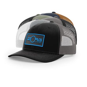 May include: A collection of trucker hats in various colors, including black, gray, olive green, and navy blue. The front of the black hat features a blue rectangular patch with the text "ZORG INDUSTRIES" in white and blue. The hats have mesh backs and curved visors.