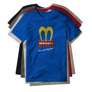 May include: A royal blue short-sleeve t-shirt featuring a yellow M logo above a red and green plaid rectangle with the white text McDowell's and the phrase Home of the Big Mick! printed below. The shirt is layered over several other t-shirts.