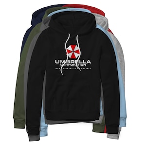 May include: A stack of hooded sweatshirts in various colors, including black, navy, and gray. The front of the black hoodie displays the Umbrella Corporation logo in white, with the text "OUR BUSINESS IS LIFE ITSELF" below.
