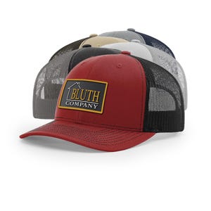 May include: A collection of trucker hats in various colors, including red, gray, and tan. The front-most hat is red with a black mesh back and a rectangular patch that reads "BLUTH COMPANY" in gold lettering.