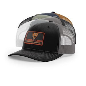 May include: A stack of trucker hats in various colors, including black, gray, and olive green. Each hat features a mesh back and a front patch with the text "TYRELL CORP MORE HUMAN THAN HUMAN" and a logo.