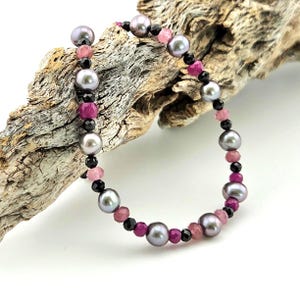 May include: A beaded bracelet featuring gray pearls, black beads, and pink and magenta gemstones. The bracelet is strung together, creating a delicate and elegant piece of jewelry. The beads are arranged in a pattern, adding visual interest.