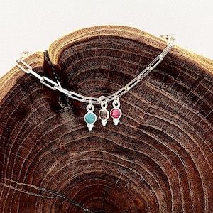 May include: A silver chain bracelet with three charms, each featuring a different colored gemstone. The charms are a teal blue, a gray, and a pink.