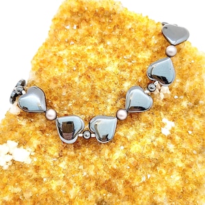 May include: A bracelet featuring heart-shaped hematite beads and small, round, pink beads. The bracelet is displayed on a textured, yellow-orange surface. The hearts have a metallic, reflective appearance.