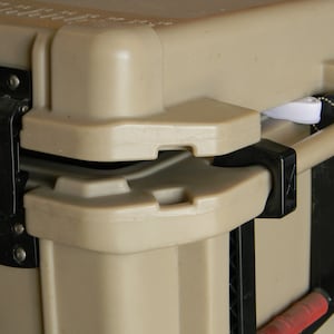 Puede incluir: Primer plano de una nevera de color beige con un cierre y un asa negros. La nevera tiene un botón rojo y un cierre blanco en la tapa.