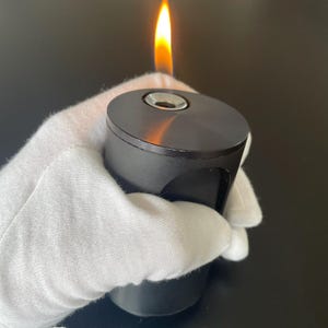 May include: A black cylindrical lighter with a flame. The lighter has a metallic finish and a small opening at the top. The flame is yellow and orange, and the lighter is held by a gloved hand.