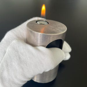 May include: A silver-colored lighter with a flame burning at the top. The lighter has a cylindrical body with a rounded top and a curved cutout on the side. The lighter is held by a gloved hand.