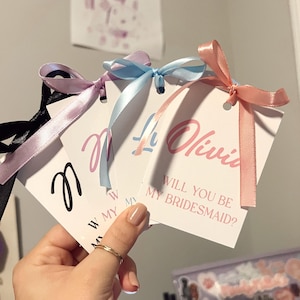 May include: Three white cards with pink, blue, and black ribbon tied around them. The cards have the text "Will you be my bridesmaid?" written on them.
