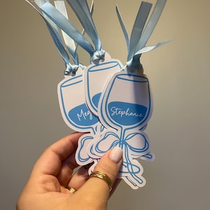 May include: Three white paper tags shaped like wine glasses with blue outlines and blue ribbon ties. Each tag features a blue graphic of a wine glass with a bow at the base and a name printed in white script, including the names Meg and Stephanie.