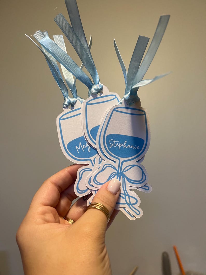 May include: Three white paper tags shaped like wine glasses with blue outlines and blue ribbon ties. Each tag features a blue graphic of a wine glass with a bow at the base and a name printed in white script, including the names Meg and Stephanie.