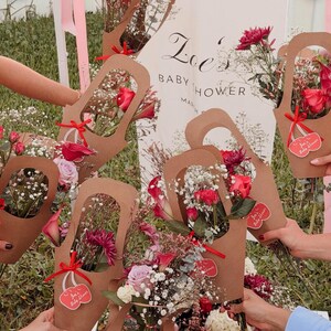 May include: Several bouquets of flowers in brown paper holders, tied with red ribbons. The bouquets contain red roses, pink and purple flowers, and baby's breath. A sign in the background reads "Zoe's Baby Shower".