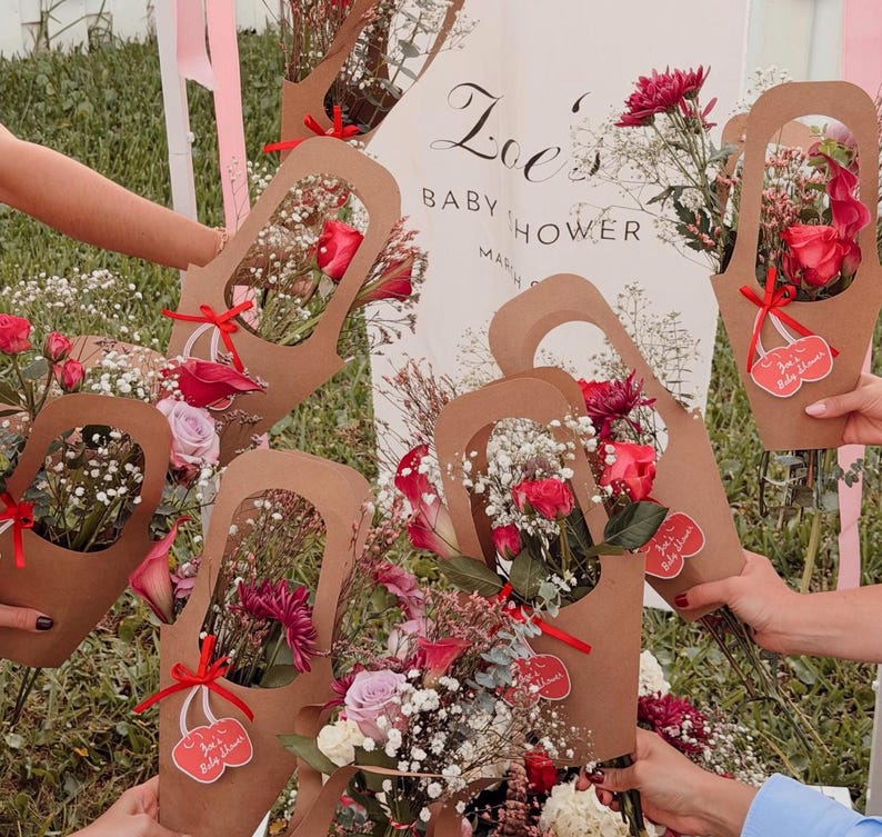 May include: Several bouquets of flowers in brown paper holders, tied with red ribbons. The bouquets contain red roses, pink and purple flowers, and baby's breath. A sign in the background reads "Zoe's Baby Shower".
